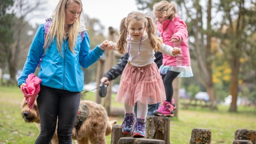 Two young girls walking along logs in the play area. A woman is holding one girl's hand and a man is holding the other girl's hand. There is also a dog on a lead.